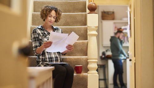a woman sits on the stairs of her home and checks a bill that she has received using her digital tablet . She is smiling happily as she checks the amounts . In the background her partner is in the kitchen .