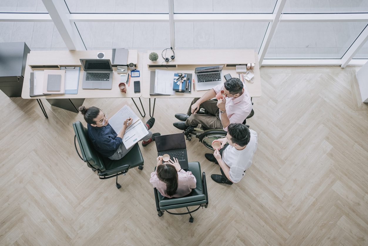 directly above Indian white collar male worker in wheelchair having cheerful discussion conversation with colleague in creative office workstation beside window